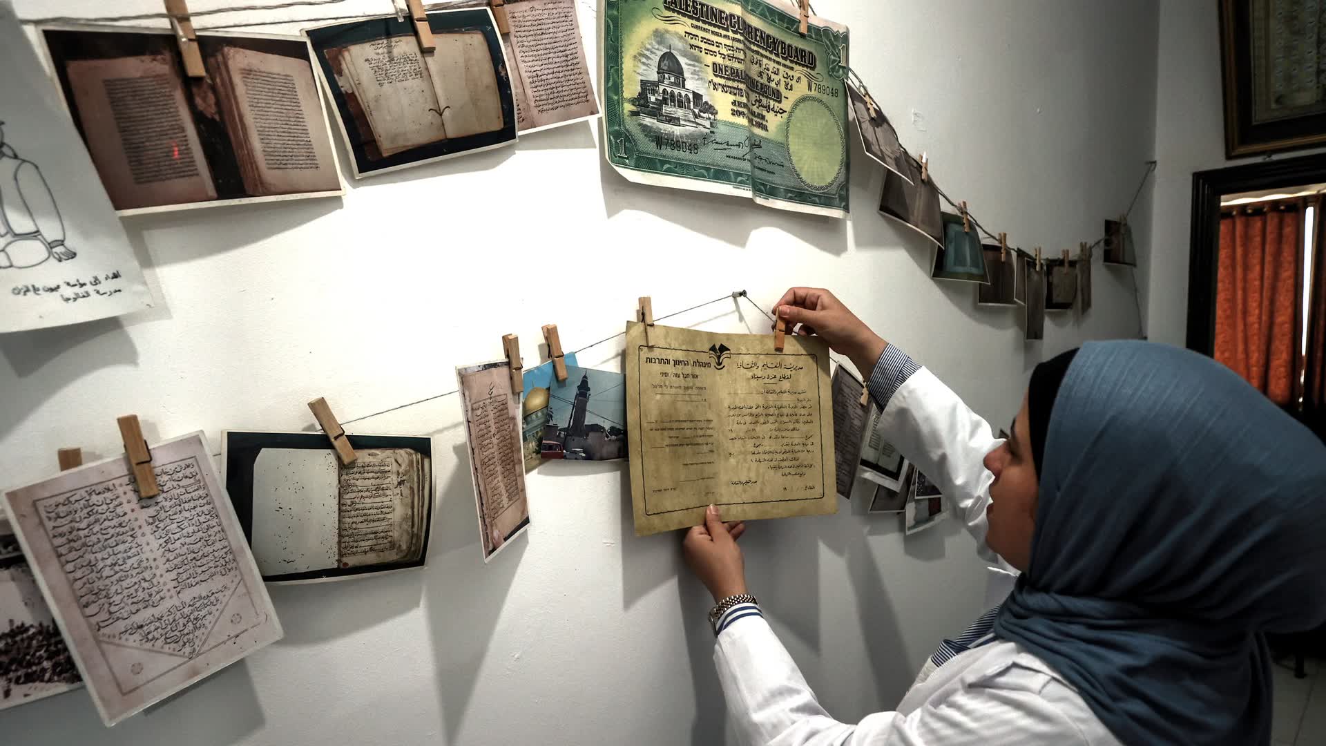 A conservator pegs a document to a drying line in a Gaza library, beside manuscript leaves and a Palestine Currency Board note pinned across the wall.