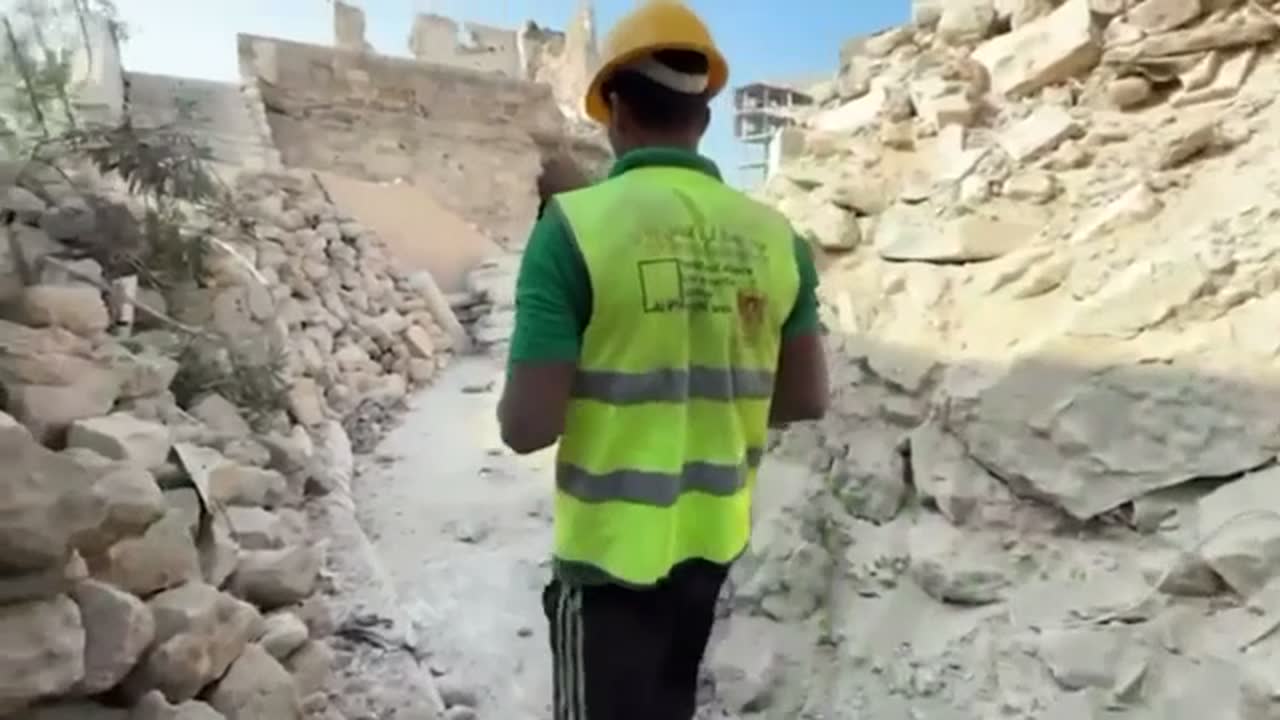 A worker crosses an open area of the Qasr al-Basha site. Cut stone is stacked at mid-frame; buildings stand behind.